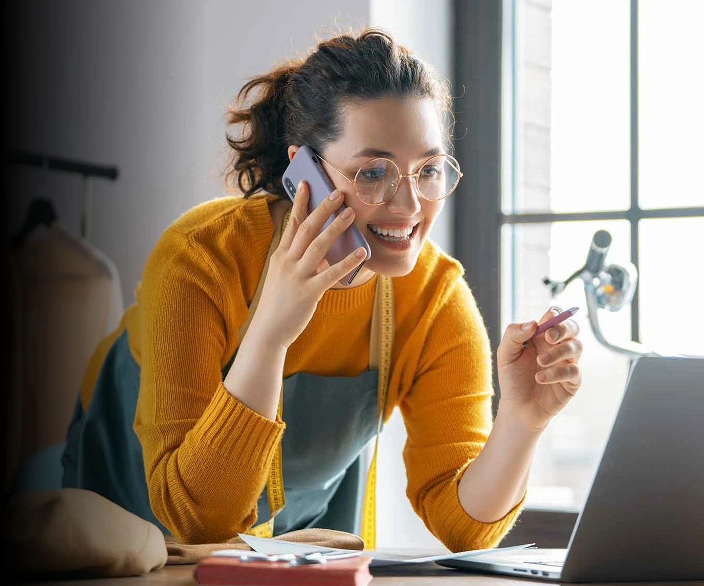 A woman in glasses looking at a laptop while holding a phone in one hand and a pen in the other